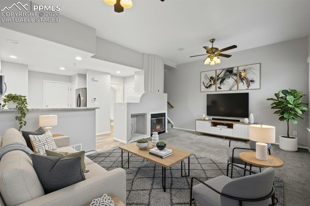 Living room featuring custom window shutters, a ceiling fan, carpet flooring, stairway, and a glass covered fireplace. This photo is virtually staged
.