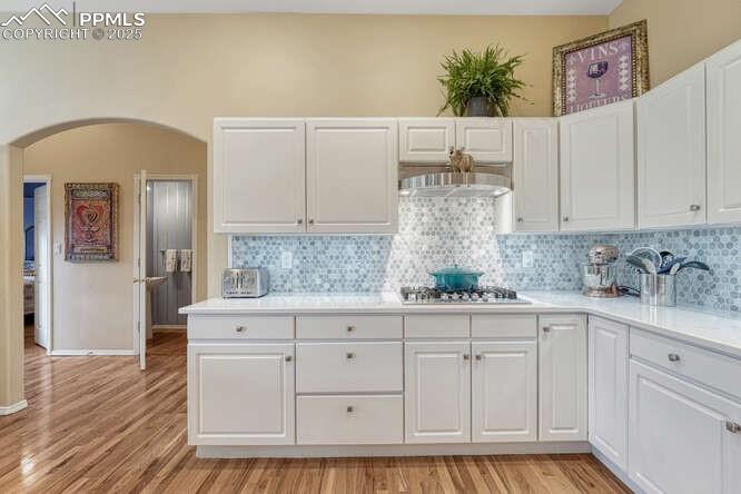 Kitchen featuring white cabinets, light wood-style floors, arched walkways, tasteful backsplash, and light stone countertops