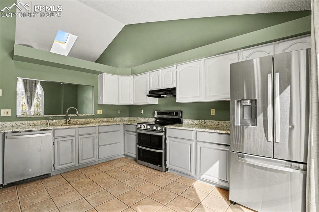 Kitchen with stainless steel appliances, light stone countertops, lofted ceiling, a skylight, and white cabinetry