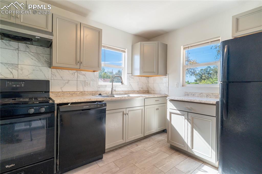 Kitchen with black appliances, tasteful backsplash, under cabinet range hood, gray cabinets, and light stone countertops
