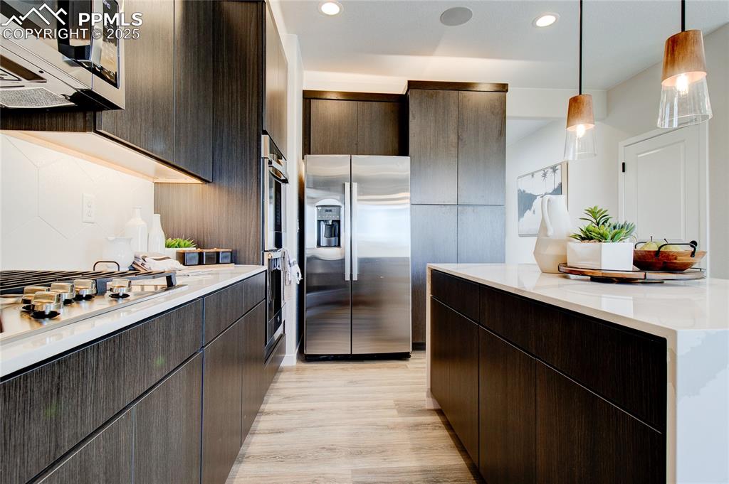 Kitchen featuring appliances with stainless steel finishes, dark brown cabinetry, light wood-type flooring, light stone counters, and hanging light fixtures