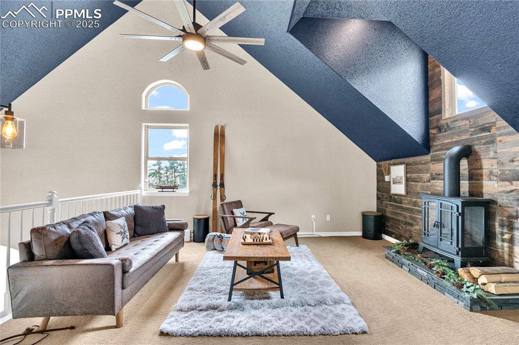 Carpeted living room featuring a wood stove, high vaulted ceiling, ceiling fan, and a textured ceiling