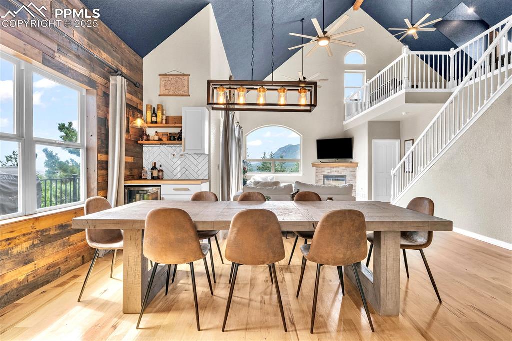 Dining room with light wood-style floors, wood walls, a fireplace, wine cooler, and high vaulted ceiling