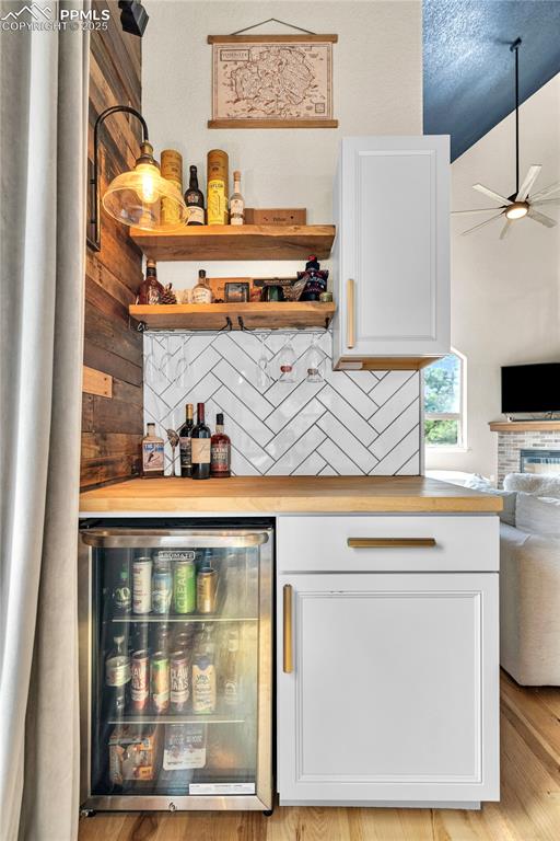 Indoor dry bar featuring beverage cooler, a fireplace, ceiling fan, backsplash, and light wood-type flooring