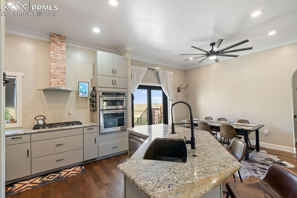 Kitchen with dark wood finished floors, crown molding, a center island with sink, appliances with stainless steel finishes, and light stone counters