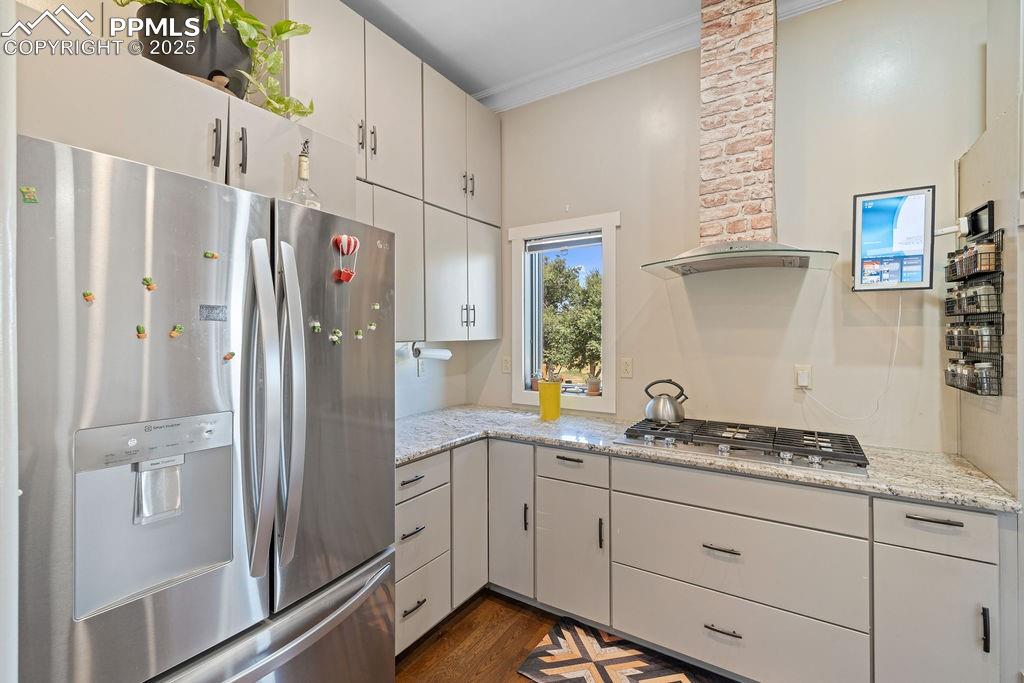 Kitchen with appliances with stainless steel finishes, wall chimney range hood, white cabinetry, dark wood-style flooring, and ornamental molding