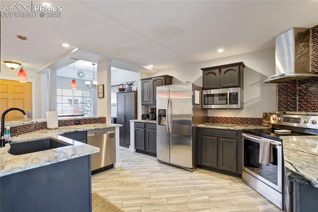 Kitchen featuring stainless steel appliances, light stone counters, backsplash, hanging light fixtures, and wood finish floors