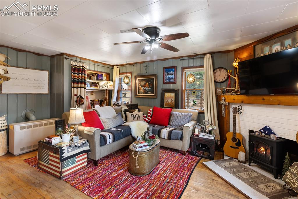 Living area featuring a wood stove, a ceiling fan, heating unit, hardwood floors, and painted wooden walls