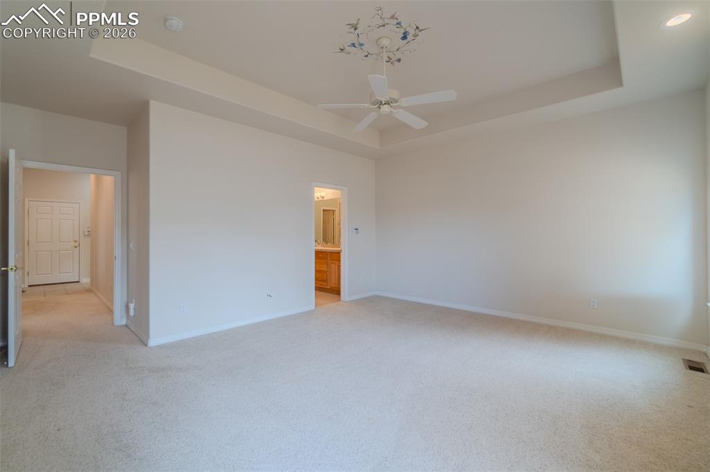 Primary bedroom on the main floor with light colored carpet, a raised ceiling, a ceiling fan, and ensuite bath