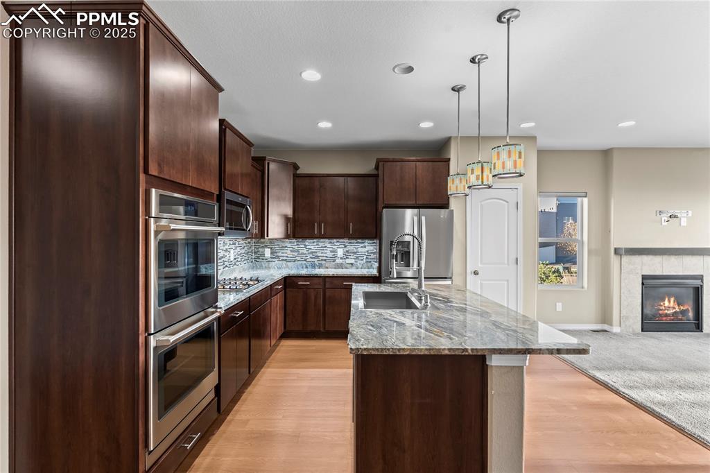 Kitchen featuring dark brown cabinets, light stone counters, a center island with sink, hanging light fixtures, and backsplash
