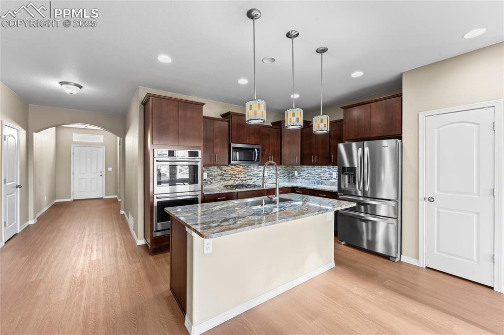 Kitchen featuring arched walkways, appliances with stainless steel finishes, dark brown cabinets, dark stone countertops, and a center island with sink