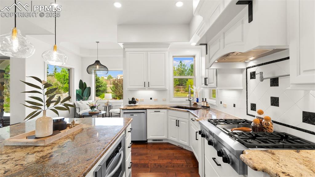 Kitchen featuring light stone counters, appliances with stainless steel finishes, premium range hood, pendant lighting, and white cabinetry