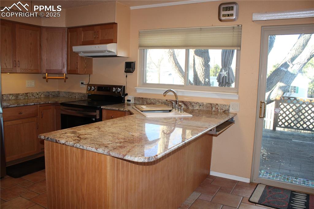 Kitchen featuring a peninsula, brown cabinetry, electric stove, and light stone counters