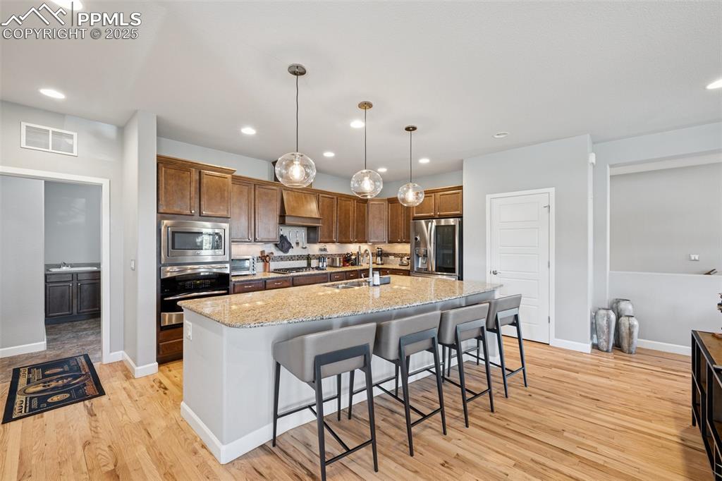 Kitchen with appliances with stainless steel finishes, custom range hood, light wood finished floors, a kitchen breakfast bar, and light stone counters