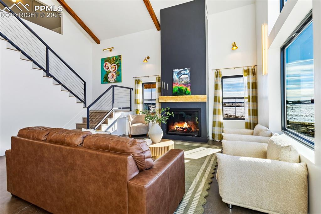 Living room featuring high vaulted ceiling, a glass covered fireplace, beamed ceiling, and stairway