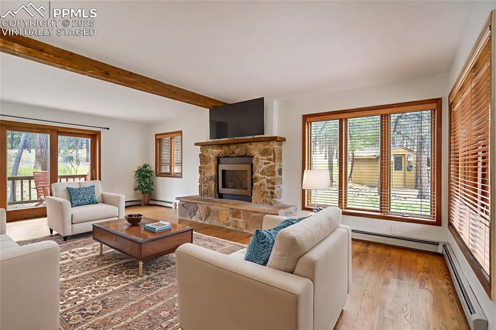 Living area featuring plenty of natural light, wood finished floors, a stone fireplace, beamed ceiling, and a baseboard heating unit, virtually staged