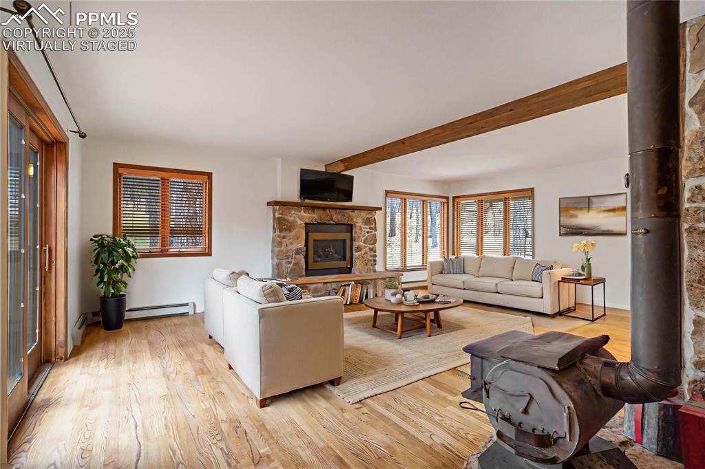 Living room with a wood stove, light wood-type flooring, beamed ceiling, and a baseboard heating unit virtually staged