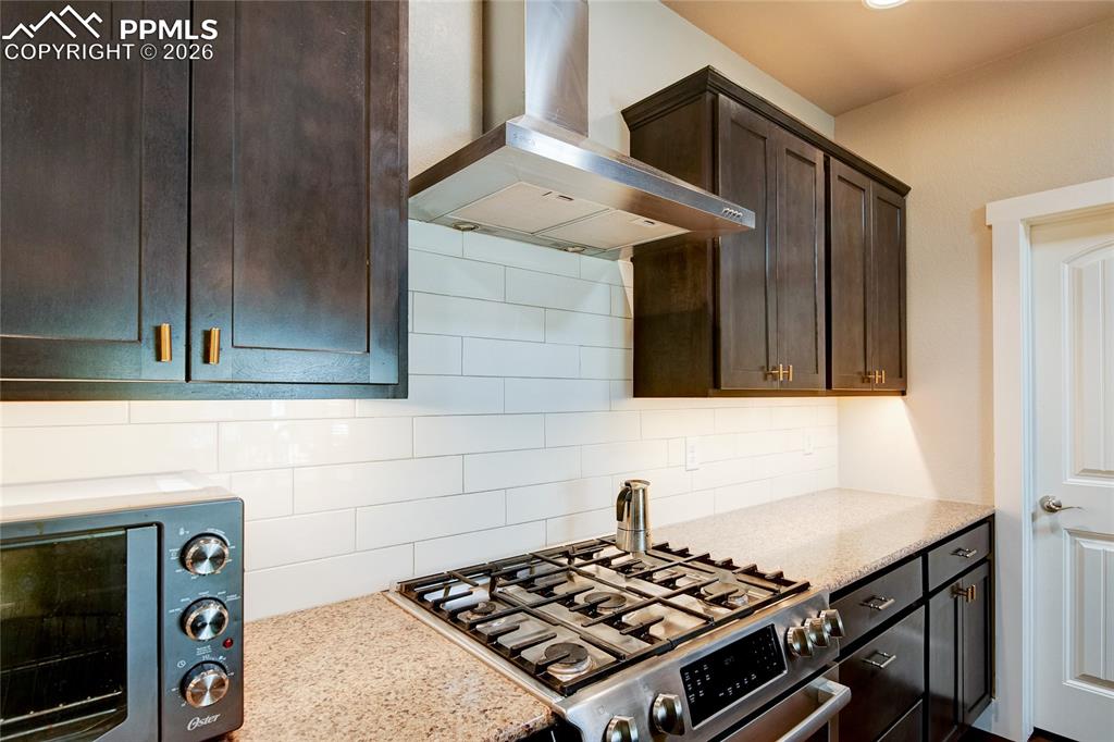 Kitchen featuring wall chimney exhaust hood, stainless steel gas range, light stone countertops, tasteful backsplash, and dark brown cabinets