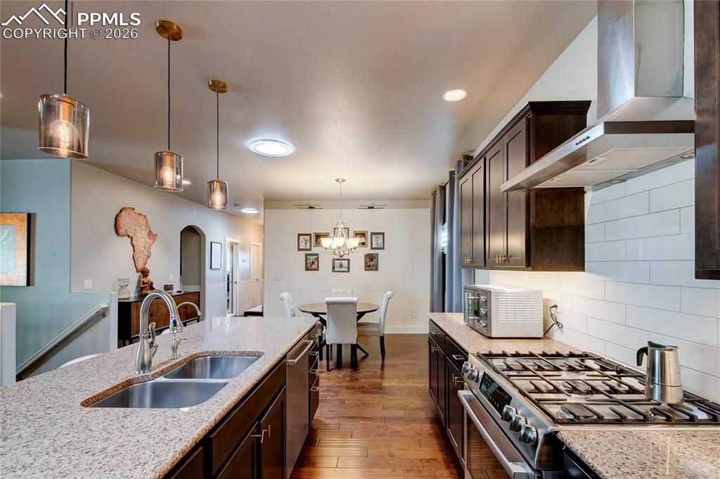 Kitchen featuring wall chimney range hood, appliances with stainless steel finishes, light stone countertops, dark brown cabinetry, and tasteful backsplash