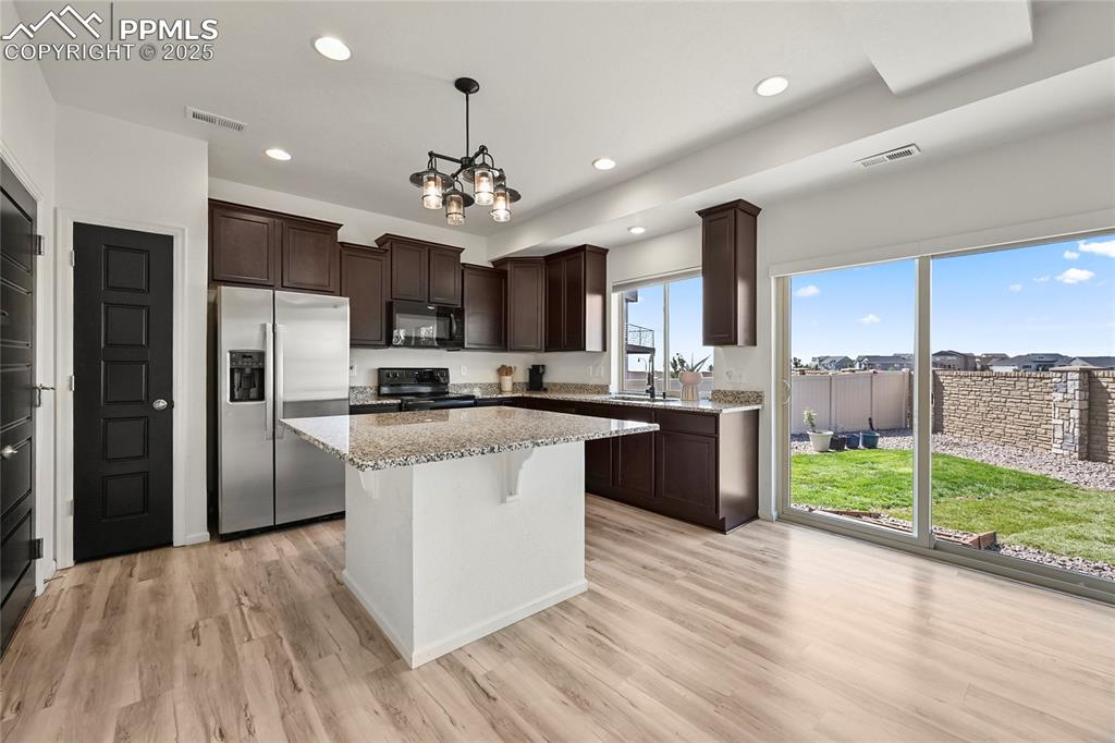 Kitchen featuring dark brown cabinetry, black appliances, light stone counters, a kitchen bar, and pendant lighting