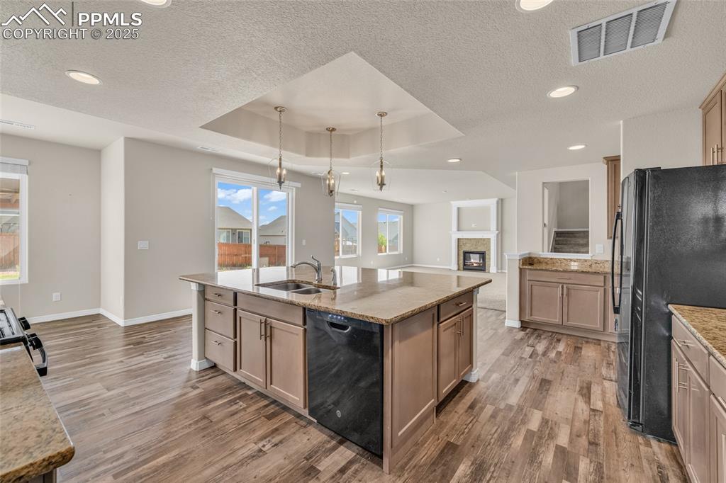Kitchen with a raised ceiling, black appliances, light wood-style floors, light stone counters, and a kitchen island with sink