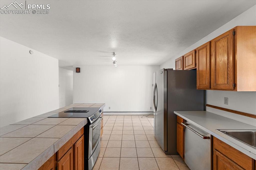 Kitchen with brown cabinetry, appliances with stainless steel finishes, light tile patterned floors, a textured ceiling, and baseboard heating