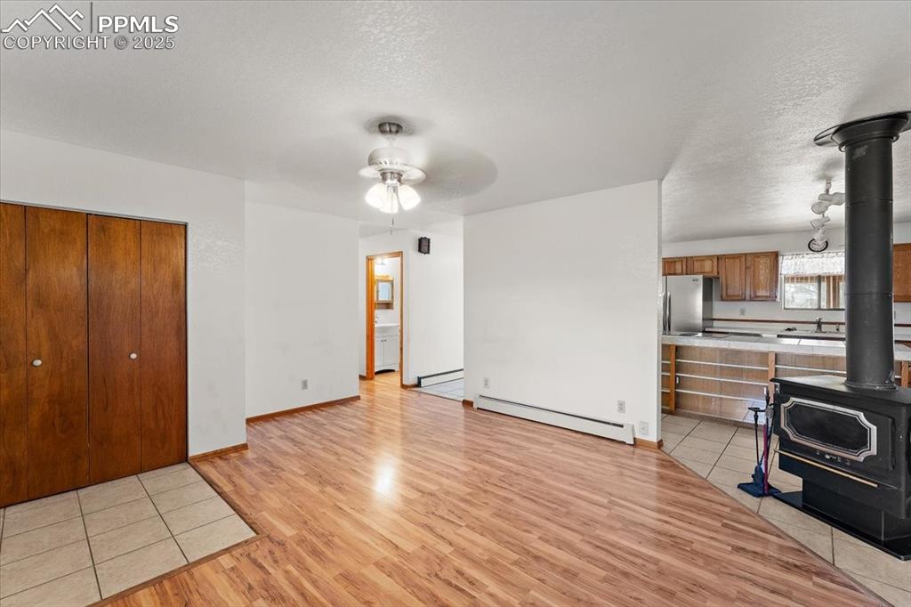 Unfurnished living room featuring a wood stove, light wood-style floors, a baseboard heating unit, a textured ceiling, and a ceiling fan