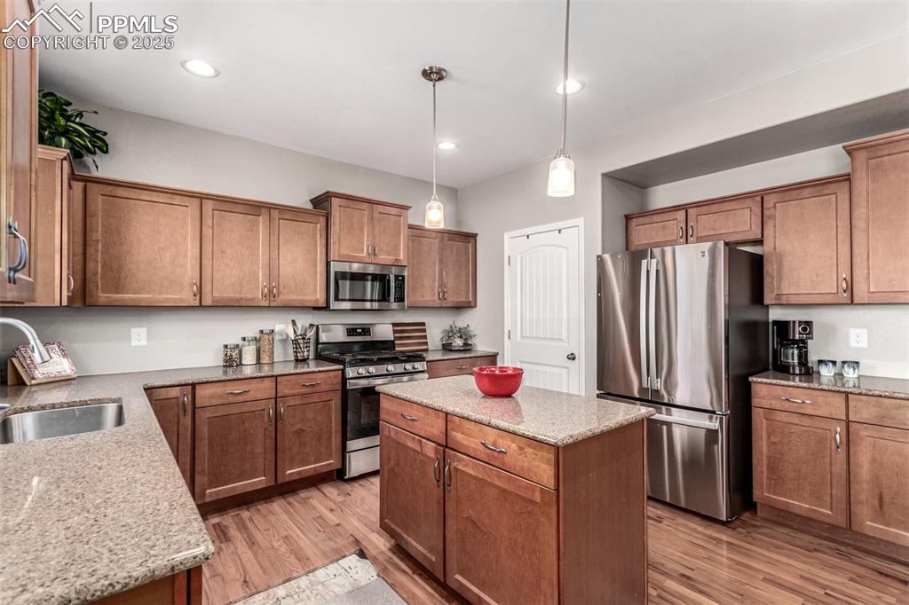 Kitchen featuring appliances with stainless steel finishes, a center island, brown cabinetry, decorative light fixtures, and light wood-type flooring