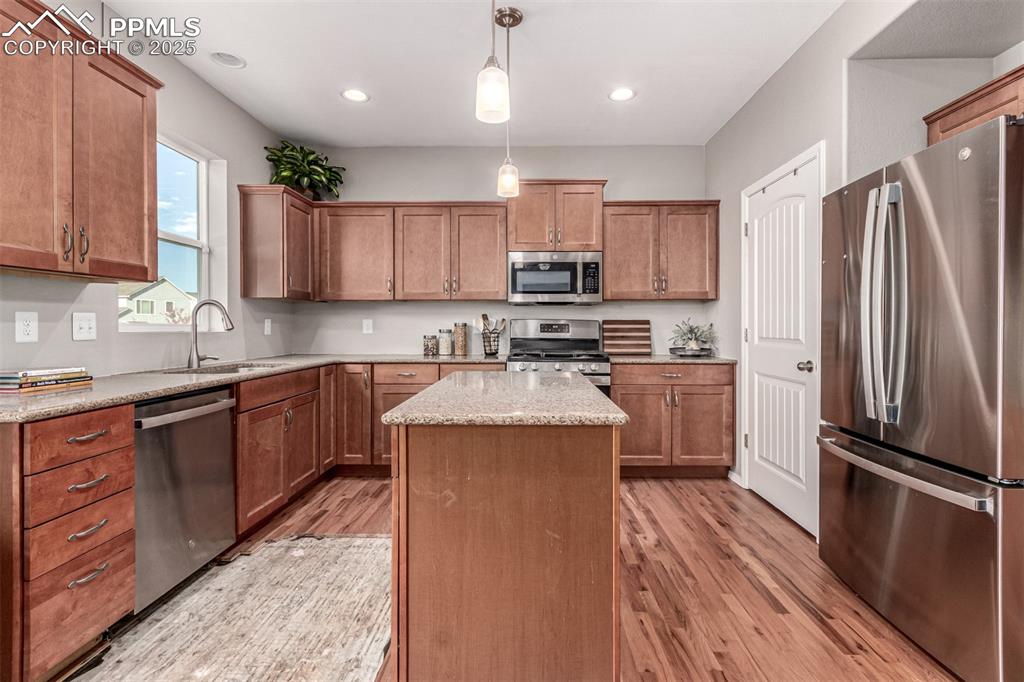 Kitchen with stainless steel appliances, brown cabinetry, decorative light fixtures, a kitchen island, and light wood-style flooring