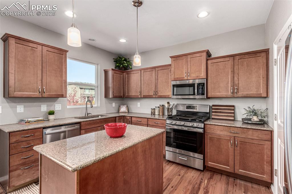 Kitchen with appliances with stainless steel finishes, brown cabinetry, a kitchen island, light wood finished floors, and decorative light fixtures