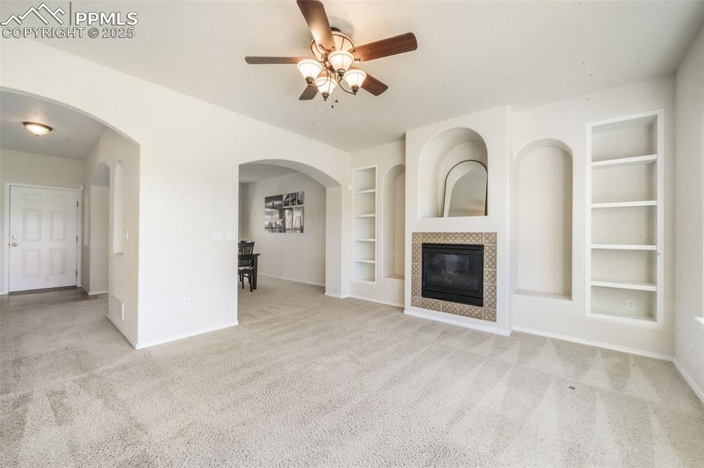 Unfurnished living room with built in features, light carpet, a tiled fireplace, ceiling fan, and arched walkways