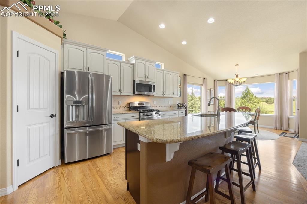 Kitchen with stainless steel appliances, a chandelier, vaulted ceiling, an island with sink, and light wood-type flooring