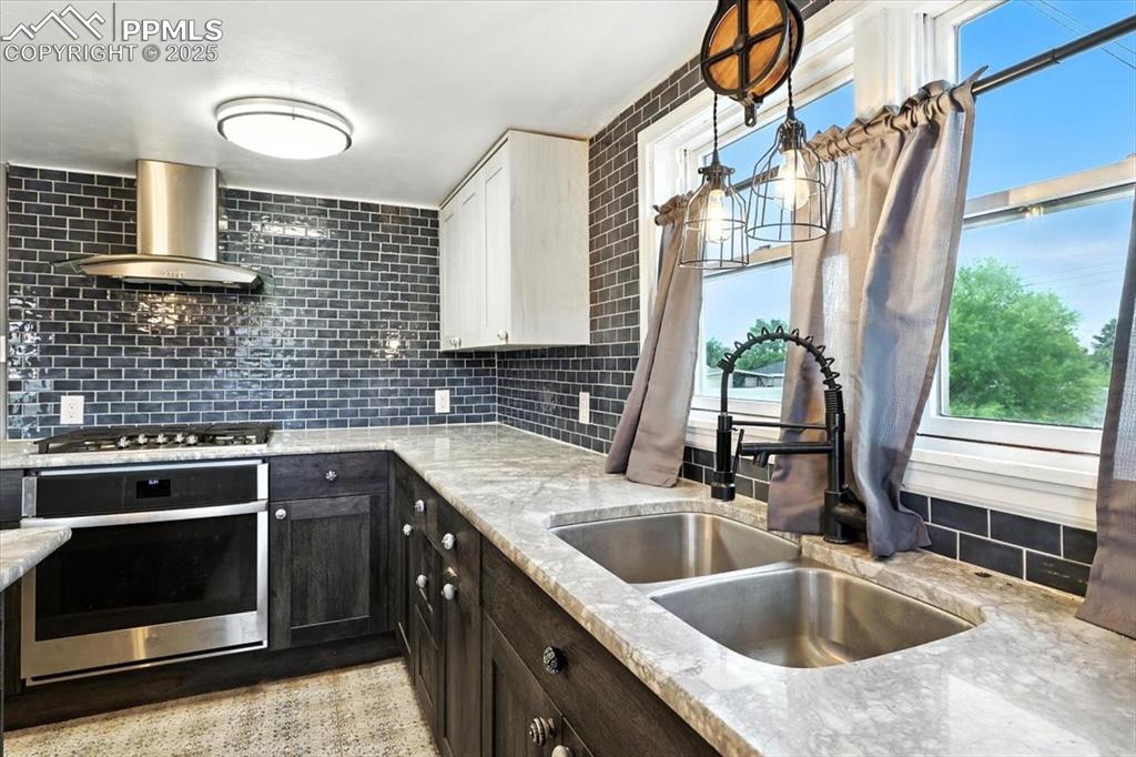Kitchen with stainless steel appliances, wall chimney exhaust hood, decorative backsplash, light stone counters, and pendant lighting