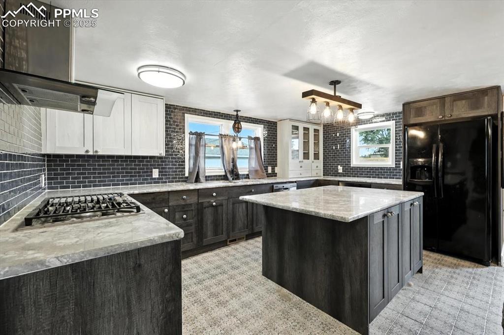 Kitchen with black refrigerator with ice dispenser, stainless steel gas cooktop, tasteful backsplash, a kitchen island, and light stone countertops