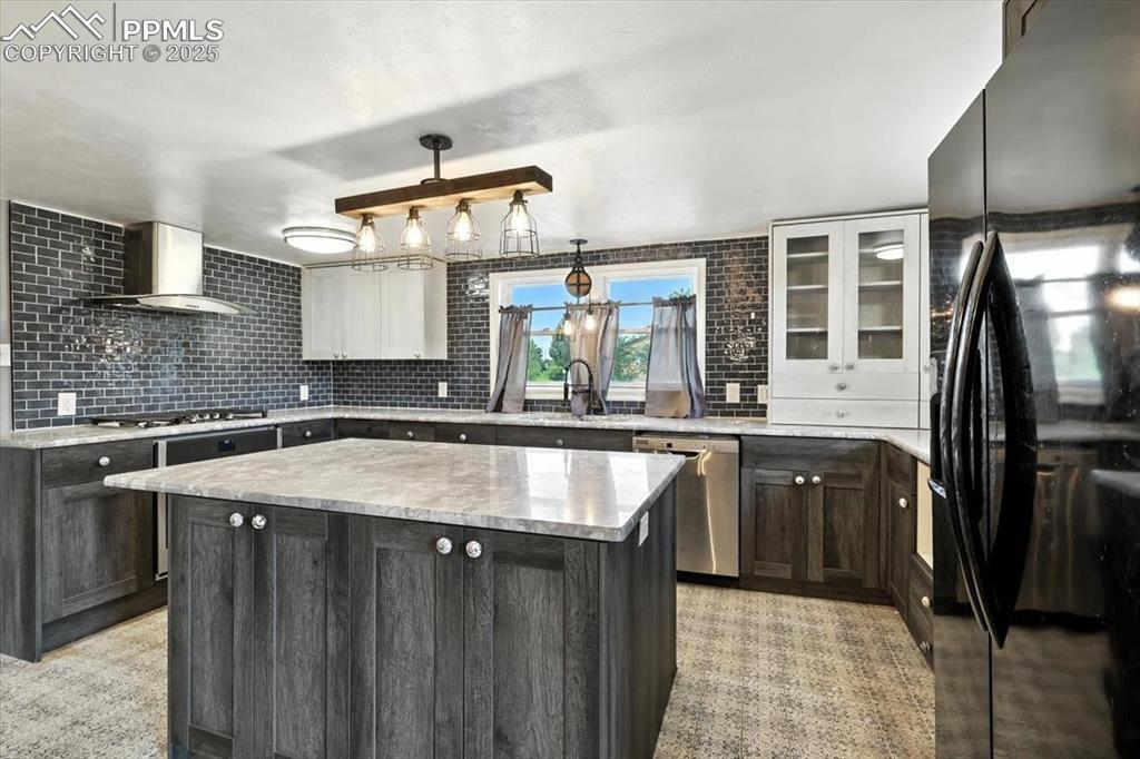 Kitchen with stainless steel appliances, wall chimney exhaust hood, a kitchen island, tasteful backsplash, and glass insert cabinets