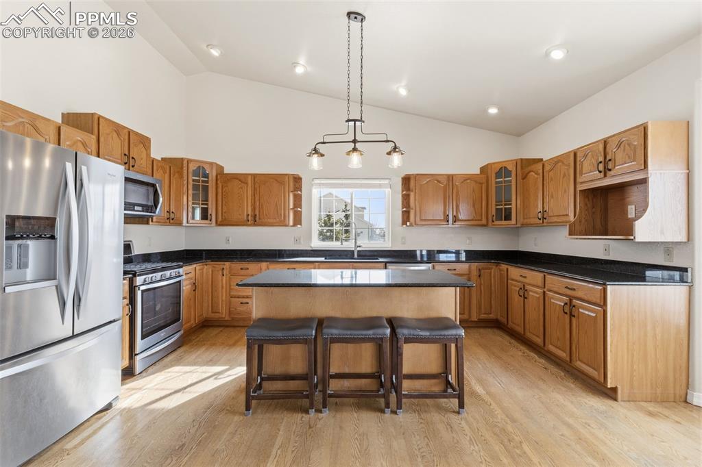 Kitchen featuring glass insert cabinets, stainless steel appliances, wood finish cabinetry, a kitchen bar, and vaulted ceiling