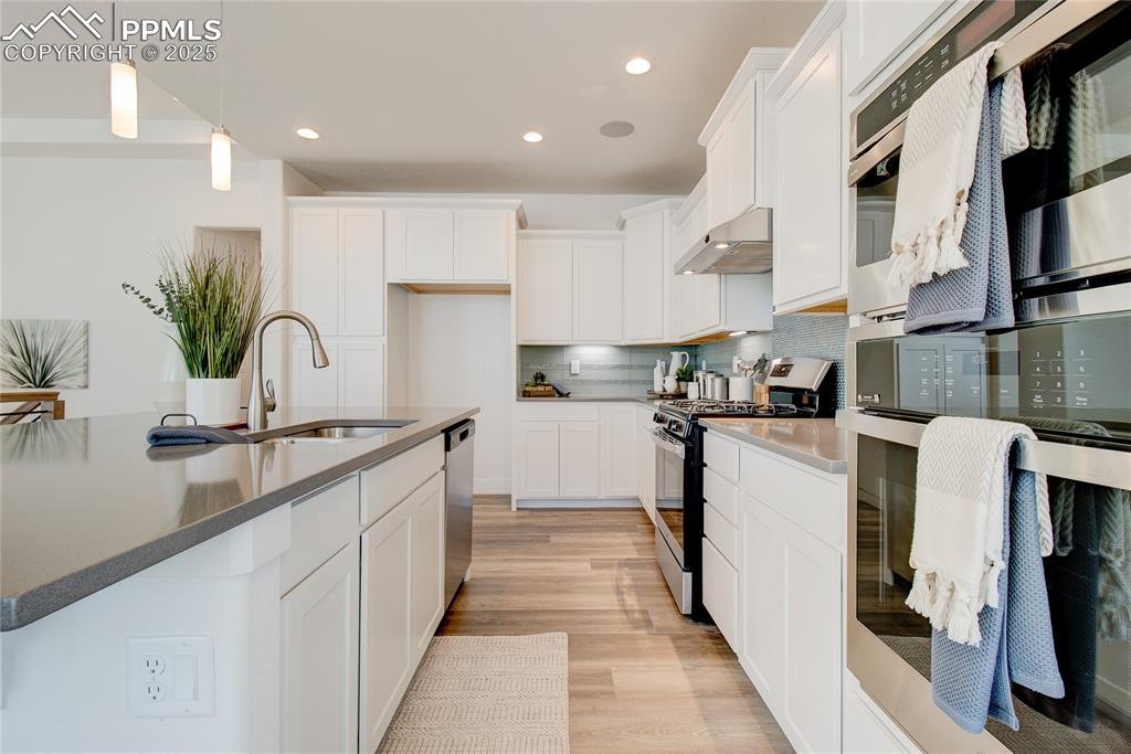 Kitchen with stainless steel appliances, under cabinet range hood, light wood-type flooring, decorative light fixtures, and white cabinets