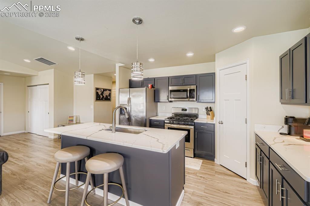 Kitchen with light stone countertops, appliances with stainless steel finishes, hanging light fixtures, an island with sink, and light wood-type flooring