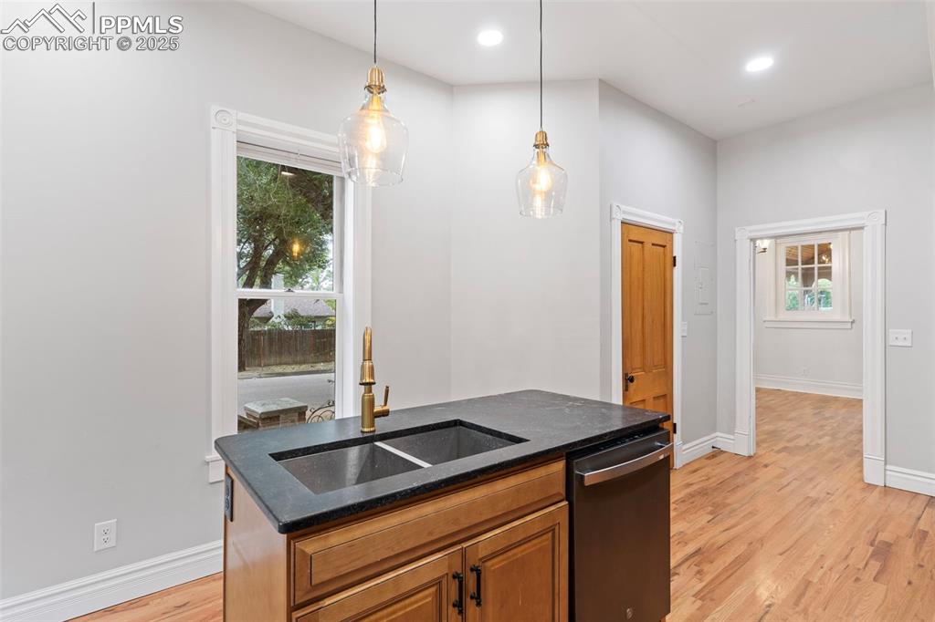 Kitchen with light wood-style floors, brown cabinets, an island with sink, dishwasher, and dark stone countertops