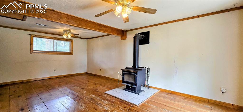 Unfurnished living room with a wood stove, wood-type flooring, beam ceiling, a ceiling fan, and crown molding