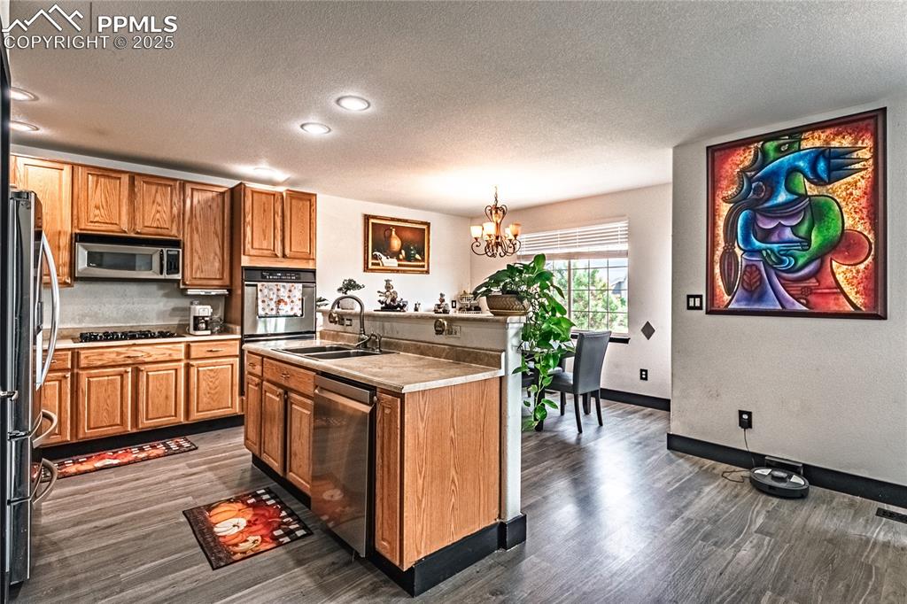 Kitchen featuring a textured ceiling, stainless steel appliances, a chandelier, brown cabinetry, and a kitchen island with sink