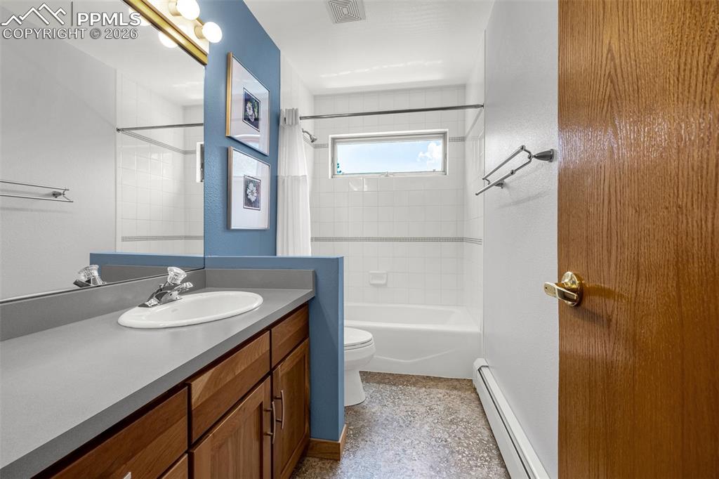 Bathroom with a baseboard radiator, vanity, shower / bathtub combination with curtain, and dark aggregate flooring