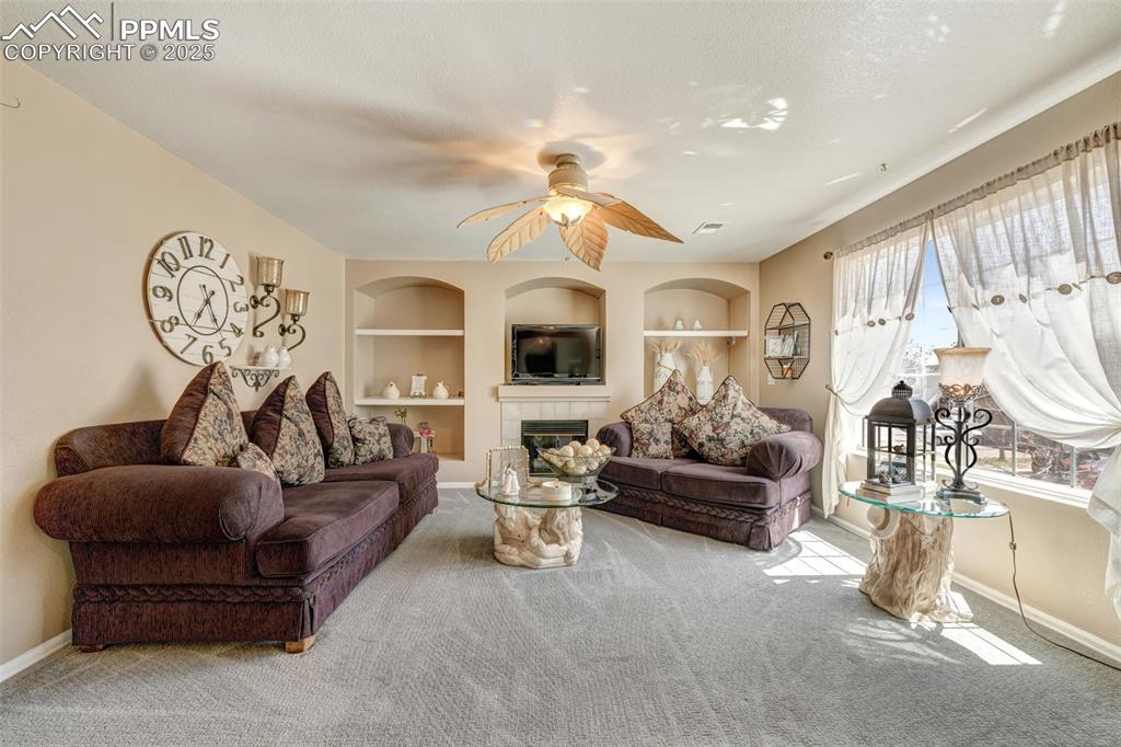 Carpeted living area featuring a tiled fireplace, built in shelves, a ceiling fan, and baseboards
