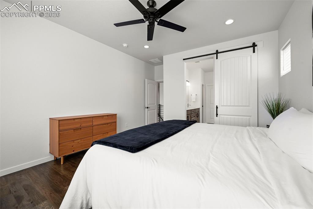 Bedroom with dark wood-type flooring, a barn door, recessed lighting, connected bathroom, and ceiling fan