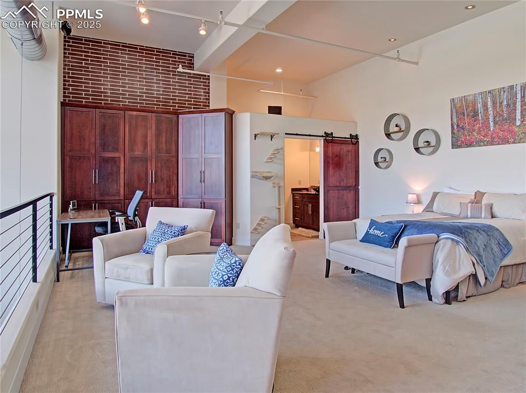 Bedroom featuring a barn door, light colored carpet, connected bathroom, brick wall, and a towering ceiling